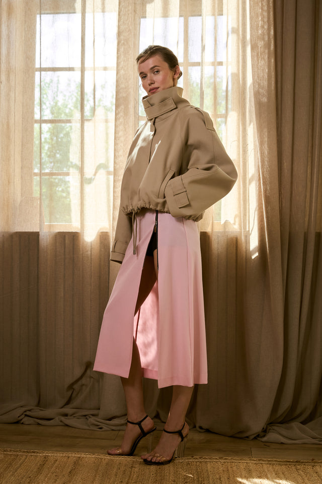 A woman standing in a room with dappled light, wearing a pink wool midi skirt with a side slit, paired with a beige top and beige ankle boots.