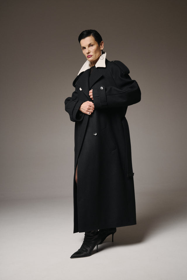 Woman wearing a long black coat with a white collar against a gray background
