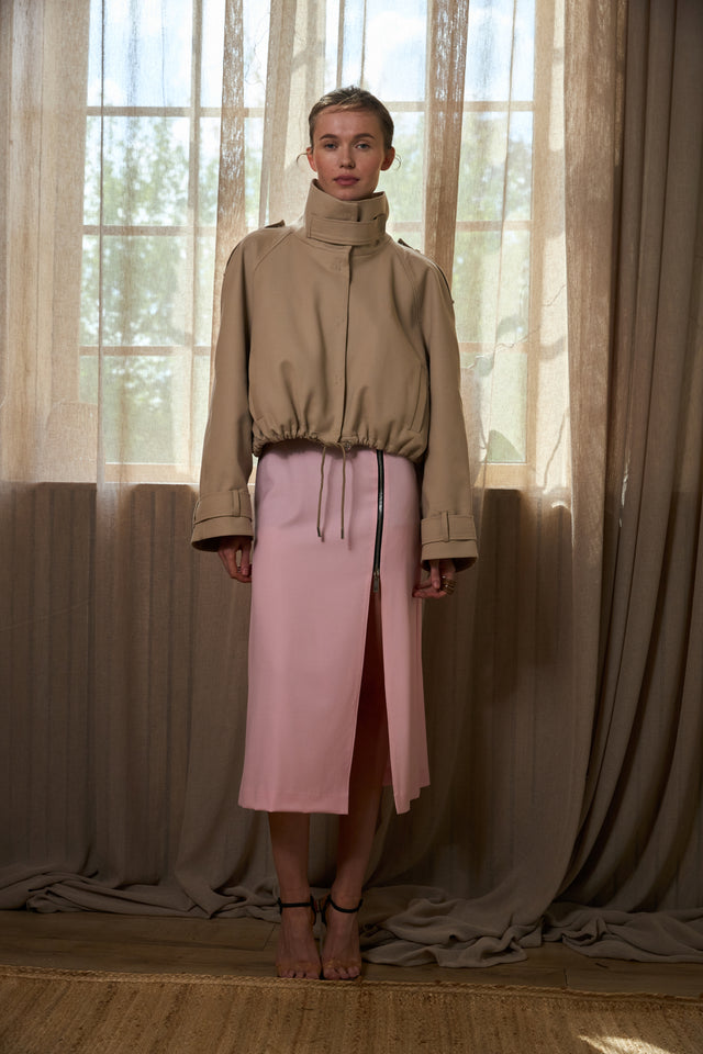 Woman in beige jacket and pink skirt standing in front of draped curtains.