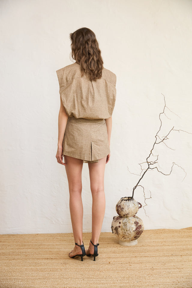 Woman in beige outfit standing with back to camera, vase on floor.
