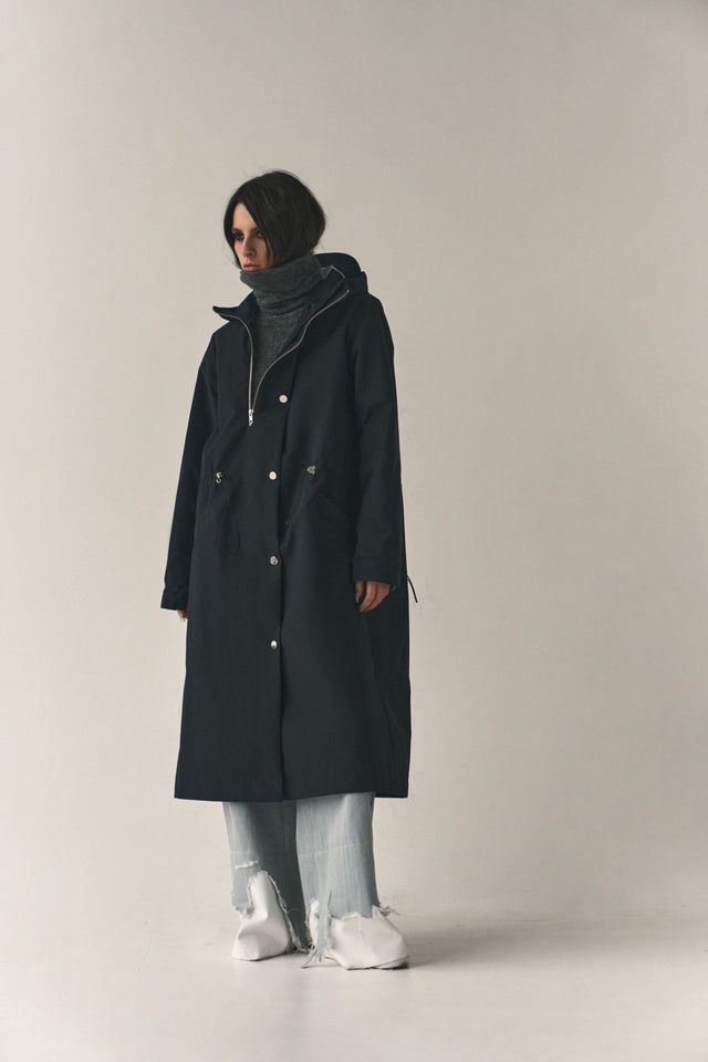 Person wearing a black coat, gray sweater, light blue jeans, and white sneakers on a plain background