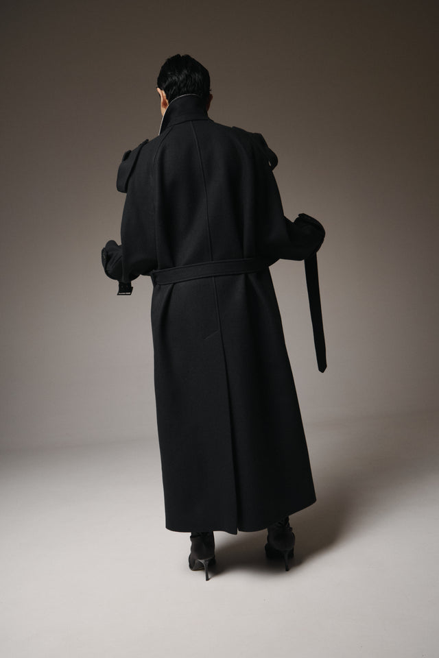 Person wearing a long black coat holding a sword, standing against a plain background