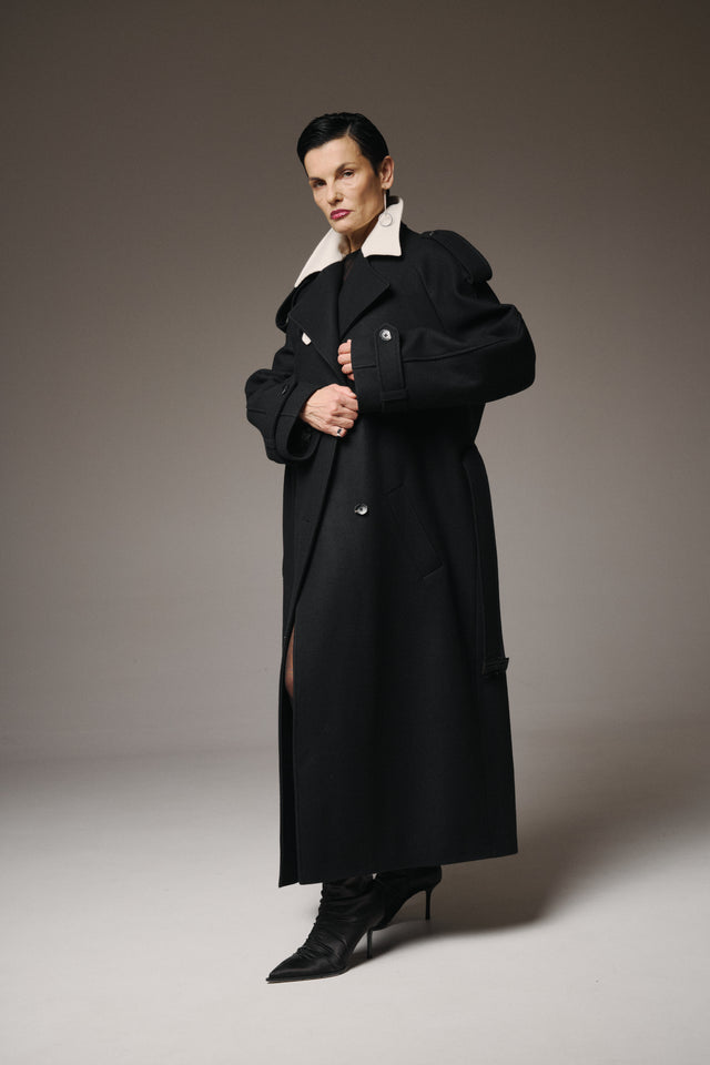 Woman wearing a long black coat with a white collar against a gray background